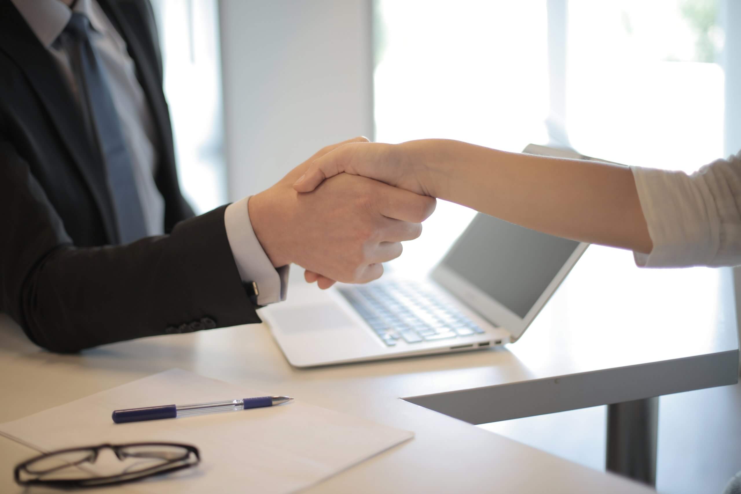 Two people shaking hands over a desk with a pen, paper, glasses, and a laptop, one person wearing a suit and the other possibly a woman.