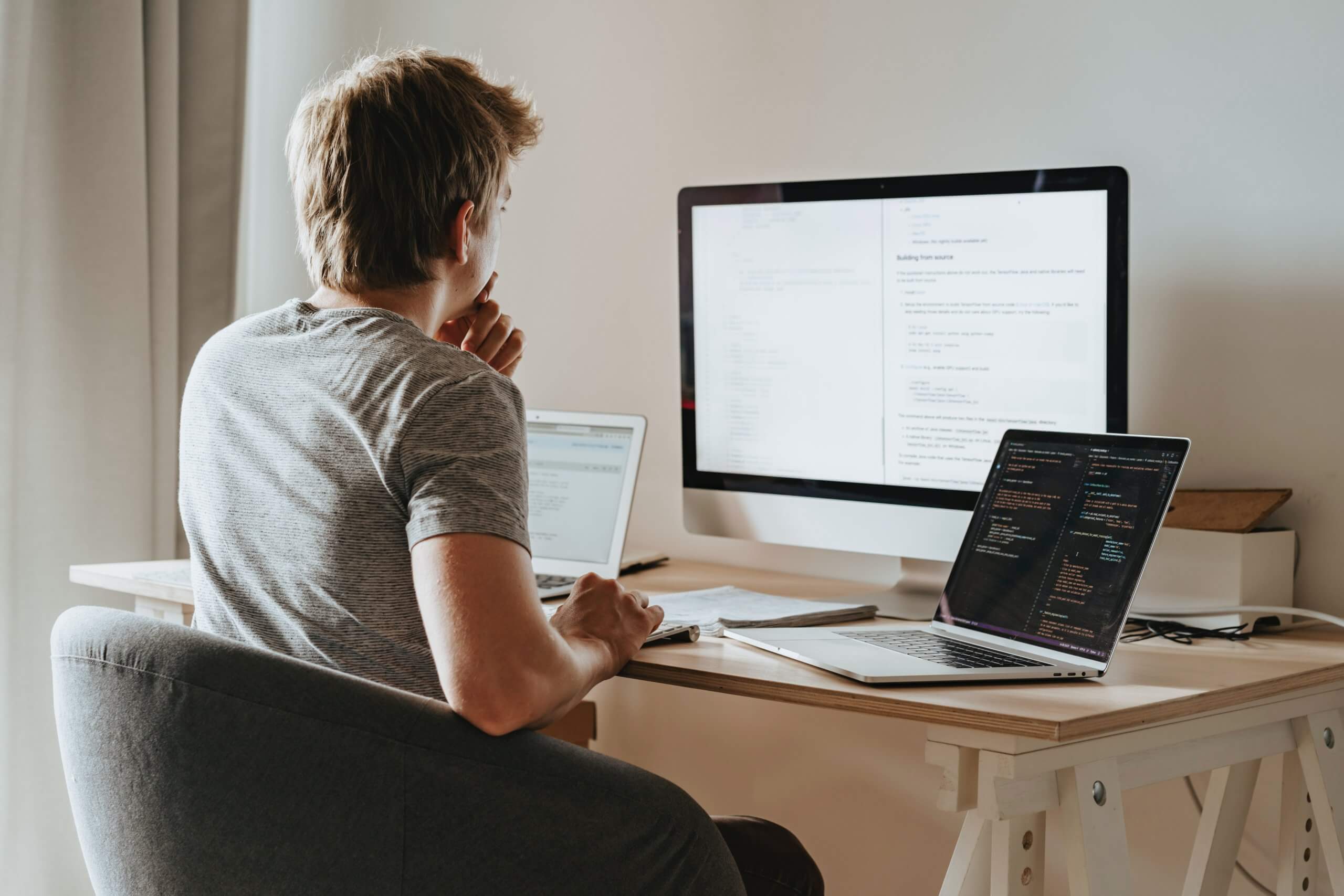 a man is sitting at his desk and looking at two laptops and one Mac coding