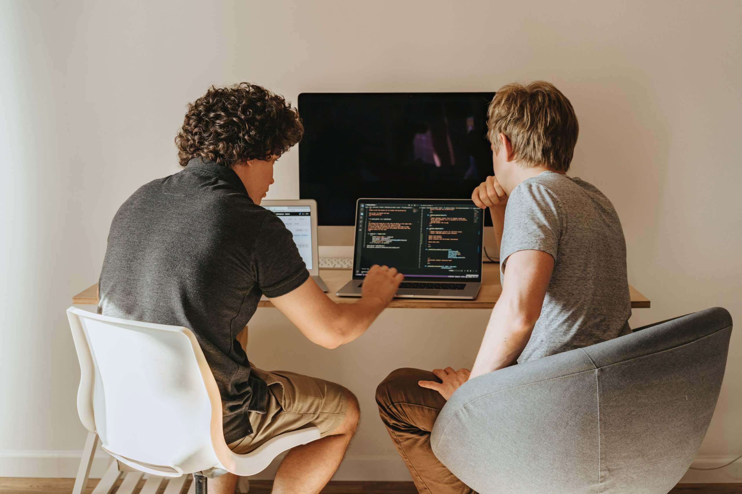 Two men sitting at one desk and looking at some code, working with two laptops and one big screen