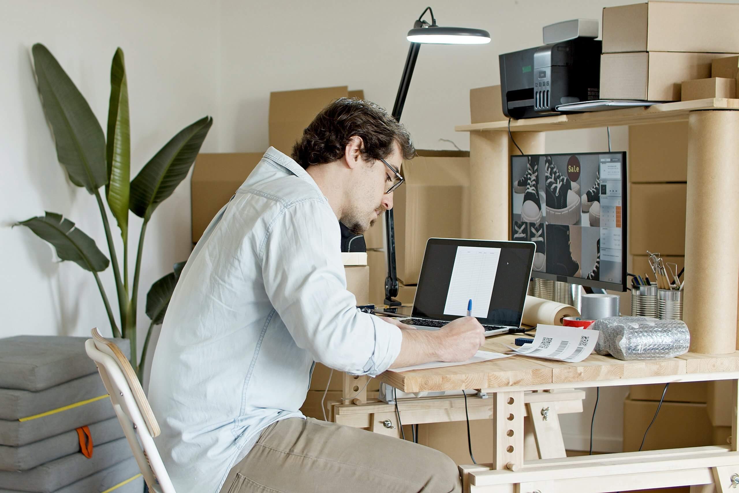 a man sitting at his desk writing notes and using a laptop, surrounded by boxes