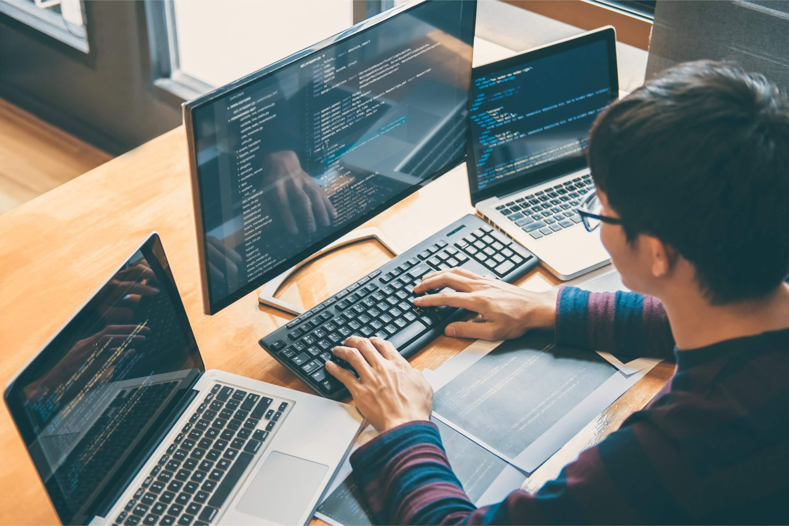 a man sitting at his desk using three screens and programming on his keyboard